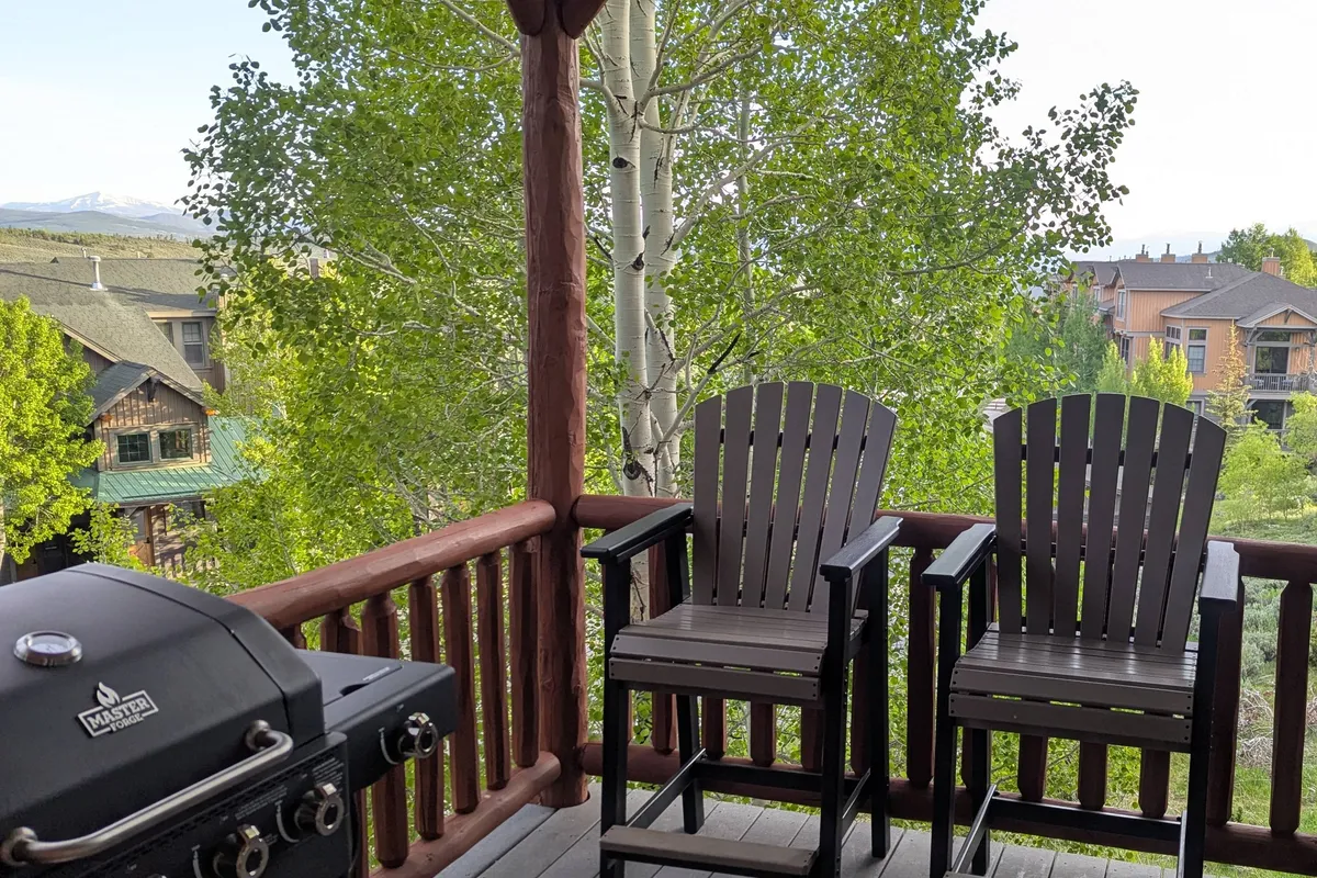 The image captures an outdoor deck area with a grill, bar stools, and a beautiful view of trees, suggesting a comfortable space for socializing and al fresco dining. The relaxed mood and amenities are ideal for summer BBQs or laid-back evenings under the open sky.