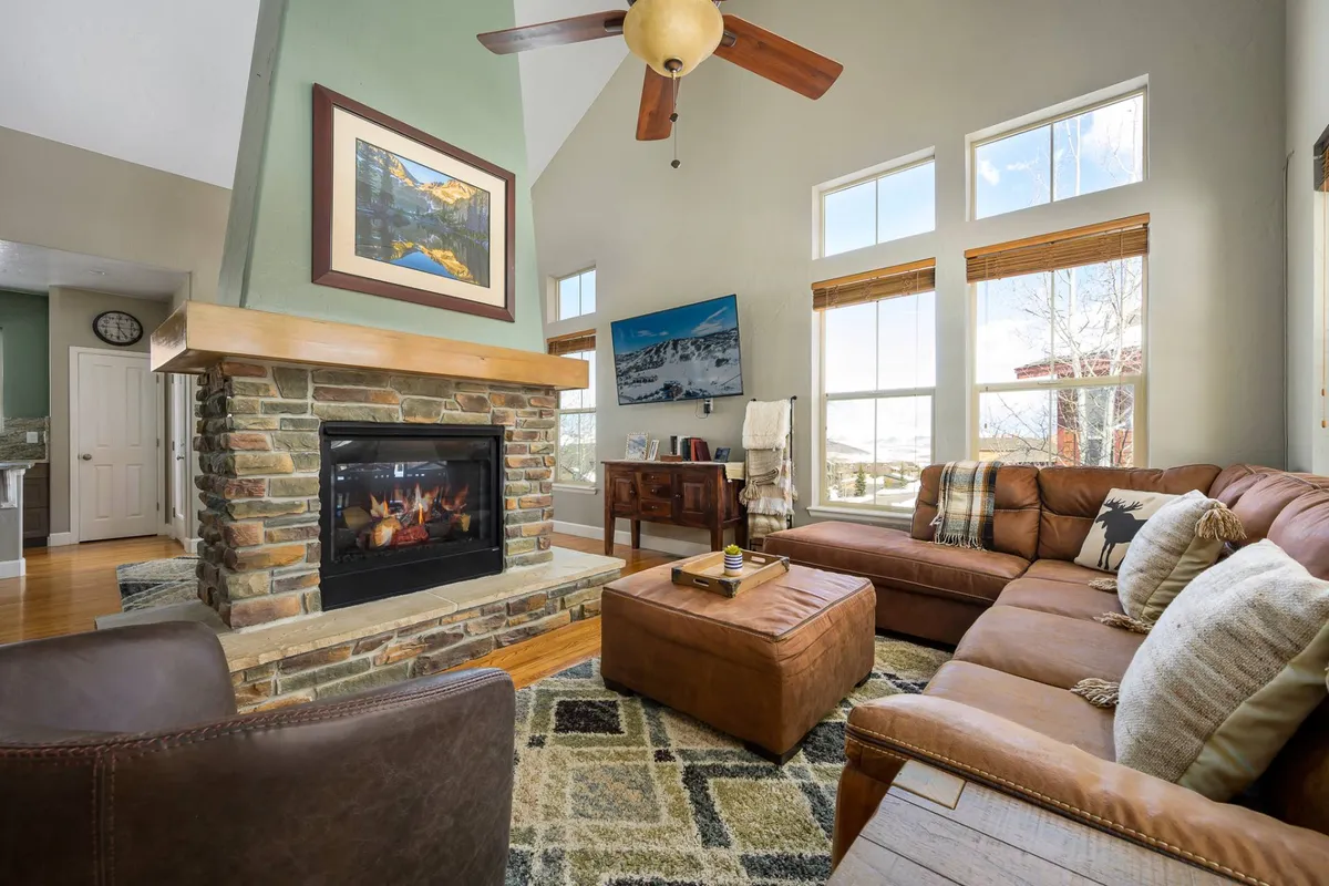 The image features a cozy interior living space with a glowing fireplace as the centerpiece, large windows offering sweeping mountain views, a plush leather sofa, and tasteful artwork. The scene captures a warm, inviting atmosphere perfect for relaxing après-ski, reading, or gathering with loved ones while enjoying the sunset through the windows.
