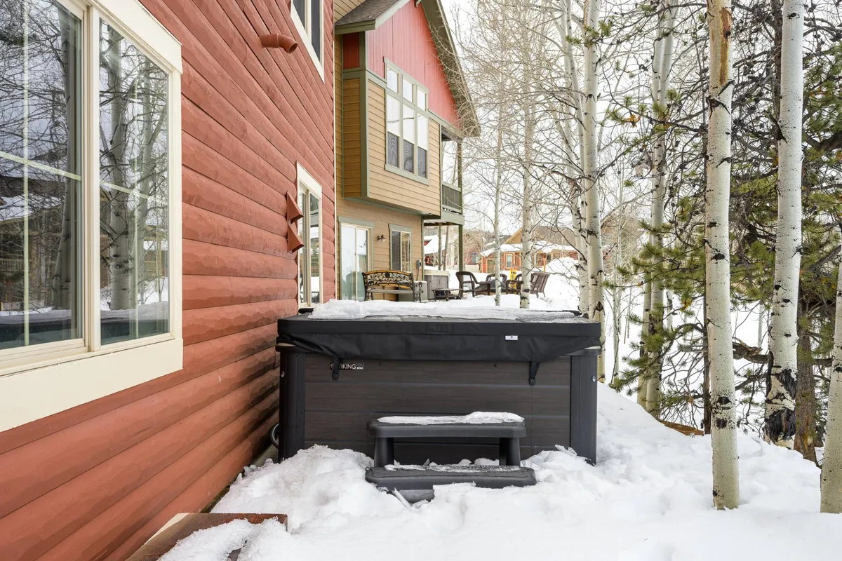 This exterior image features a private hot tub set against a snow-covered ground, bordered by aspen trees and the wooden siding of the house. The setting is serene and inviting, perfect for relaxing after a day of mountain adventure. The scene hints at stargazing opportunities and the warmth of the hot tub amid winter scenery.