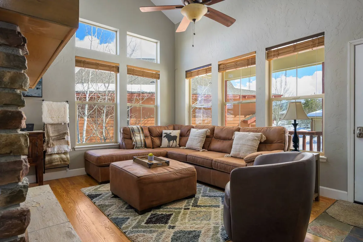 A spacious living room featuring a leather sectional sofa surrounded by large windows that invite in natural light. Decorative pillows and a coffee table create a welcoming atmosphere, while the windows provide glimpses of the mountain landscape outside.