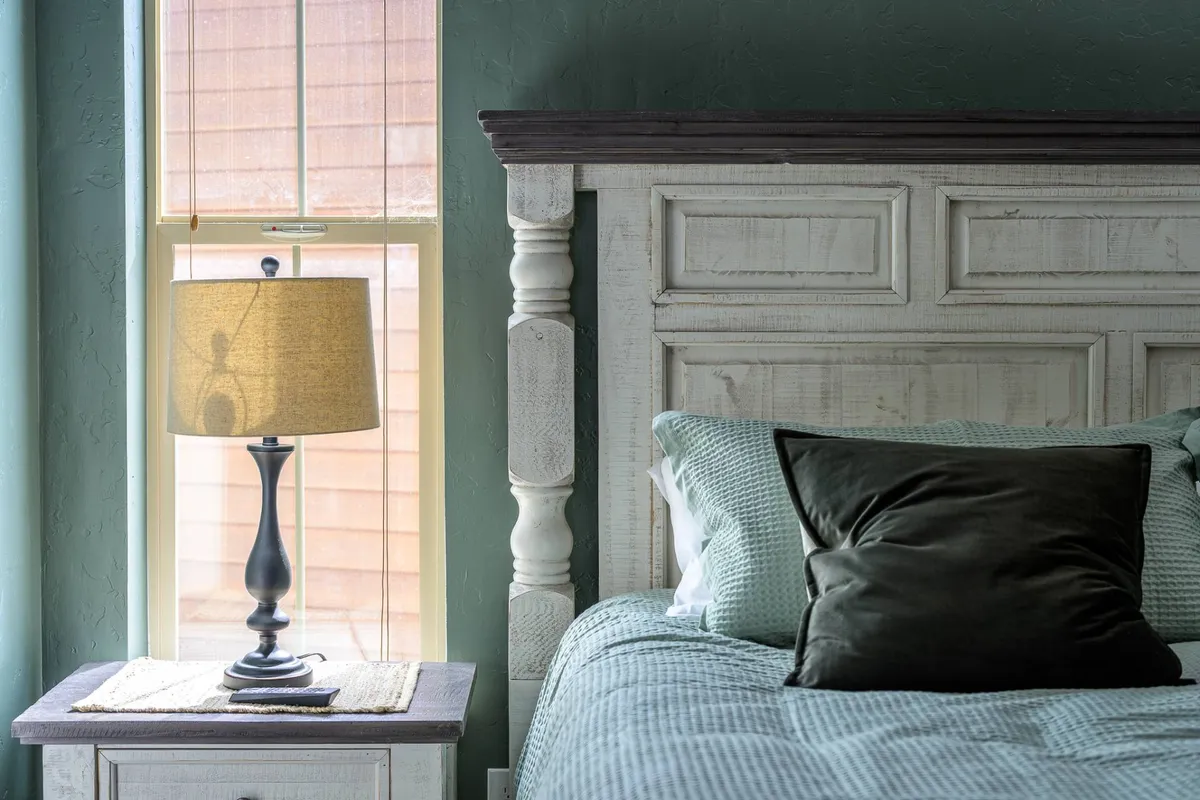 A serene bedroom featuring a plush bed with a classic headboard, soft pillows, and a lamp by the window. Natural light pours in, illuminating the tranquil space and highlighting the inviting, restful atmosphere.