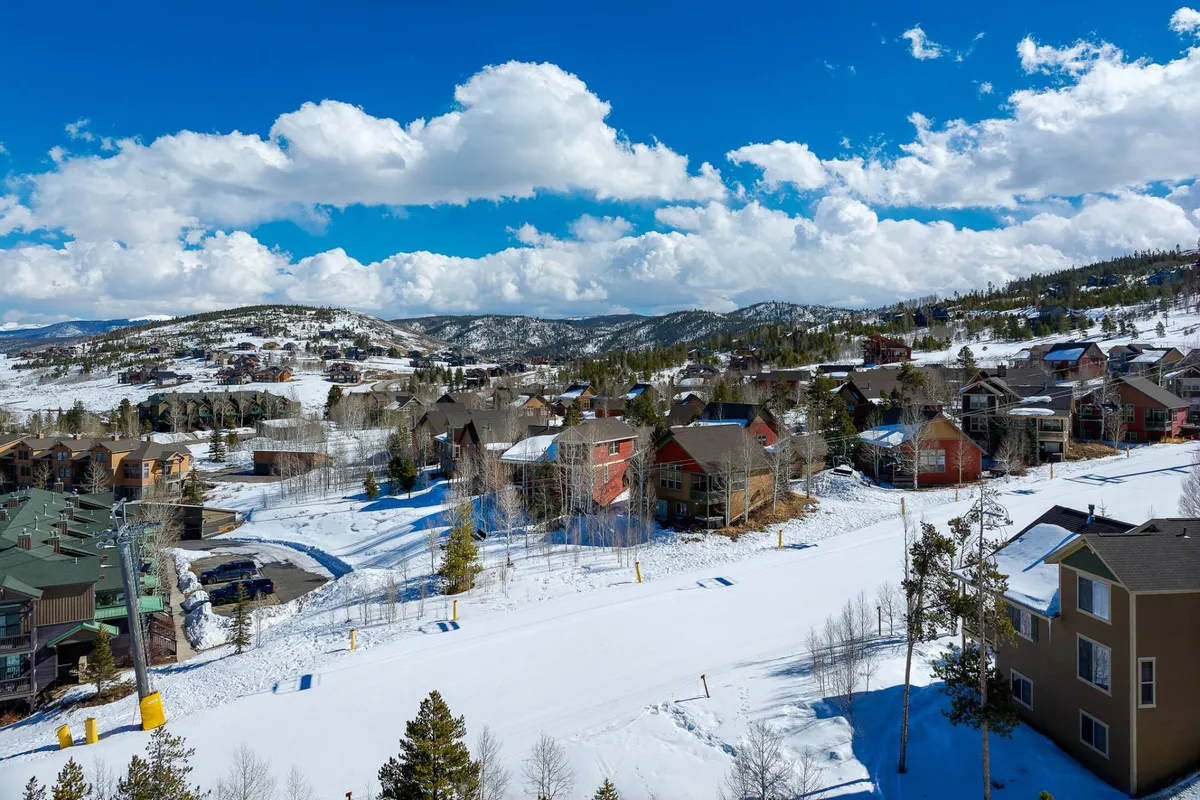 A wide view of a snow-covered landscape with colorful houses, distant mountains, and a cloudy blue sky. The scene is vibrant and expansive, featuring the neighborhood’s mountain backdrop and open spaces ideal for outdoor activities.