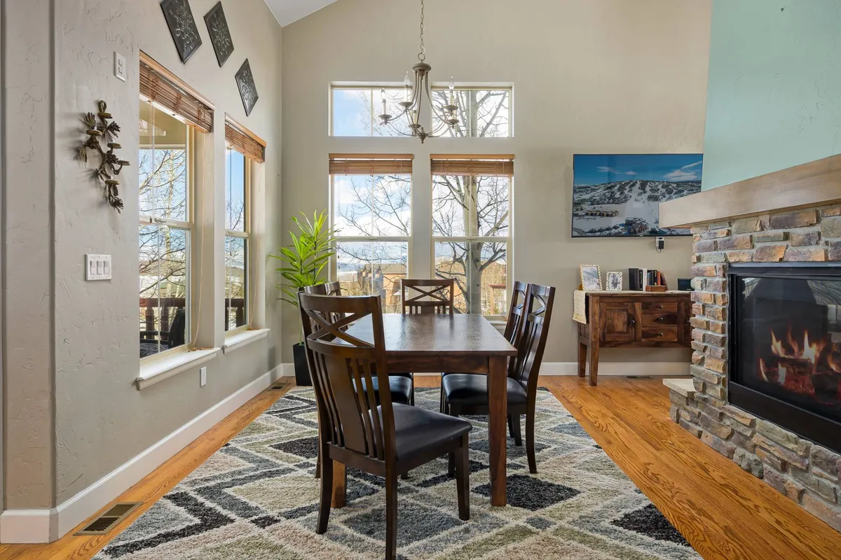 An elegant dining area anchored by a wooden table, set beside a fireplace and framed by large windows. The ambiance is warm and welcoming, ideal for family dinners or festive holiday celebrations. The expansive windows reveal views of the surrounding landscape, enhancing every meal with a scenic mountain backdrop.
