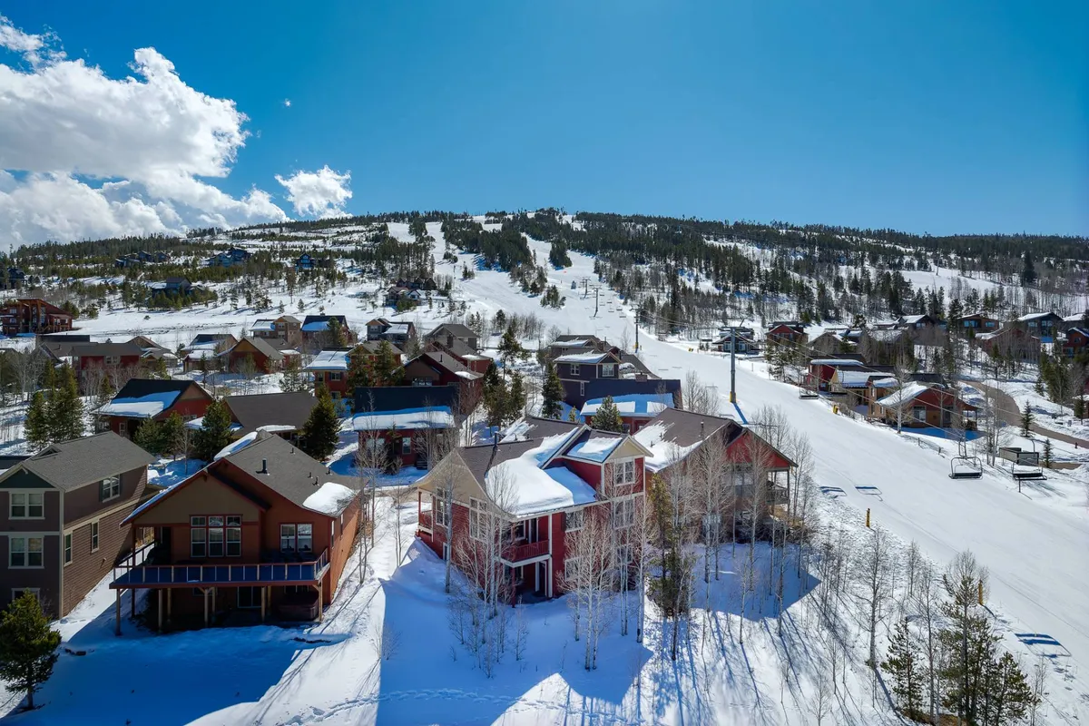 A vibrant exterior view featuring snow-covered ski slopes directly behind a cluster of colorful residential houses, framed by tall pine trees. The early morning sunlight bathes the scene, highlighting the seamless access from home to ski slopes—a true ski-in/ski-out experience. The cheerful mood captures the excitement of winter sports and the warmth of the neighborhood.