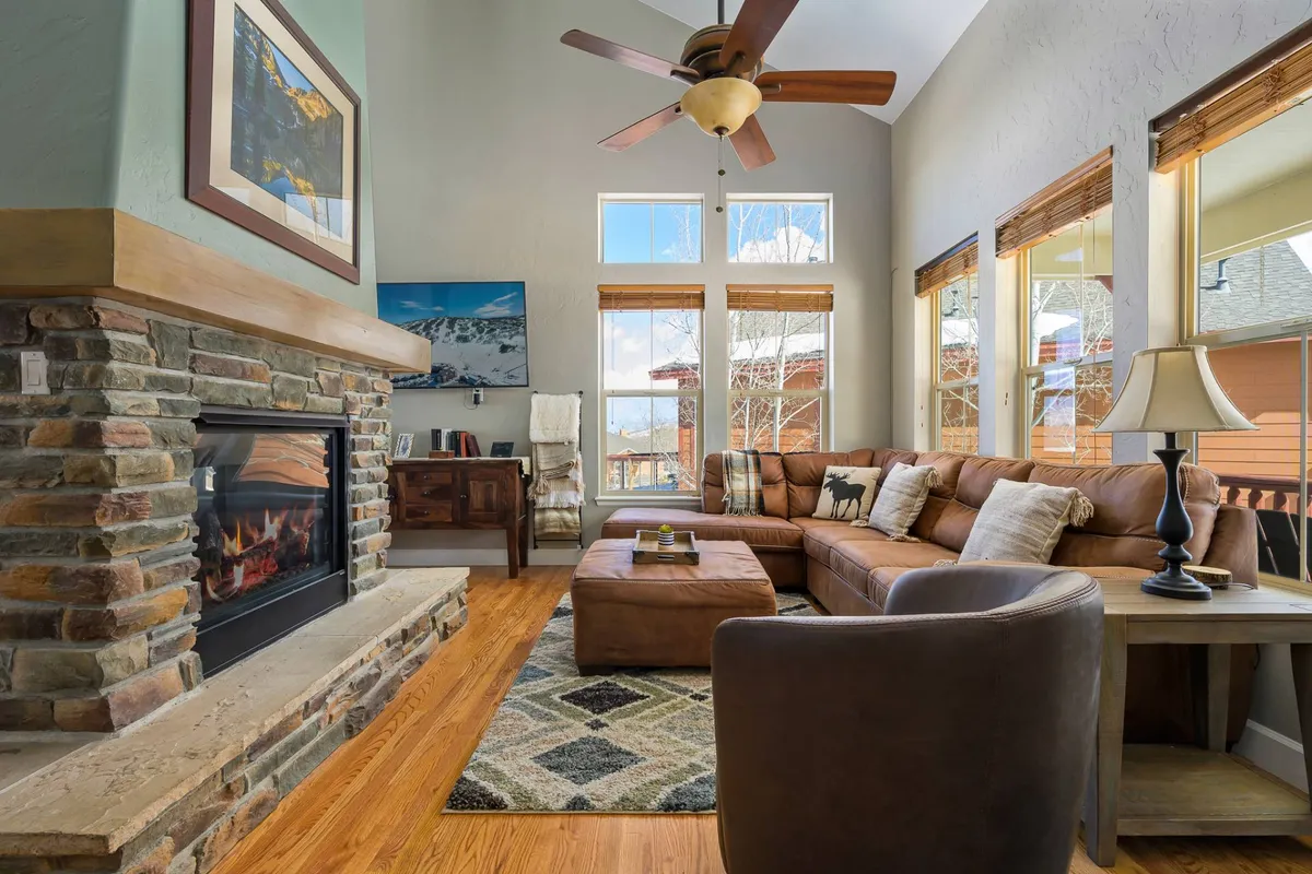 A warmly lit living area with a stone fireplace as the centerpiece. Large windows flood the room with natural light and offer mountain views. The leather sofa and wooden coffee table create a comfortable space for relaxing or socializing. The mood is welcoming and perfect for winter gatherings.