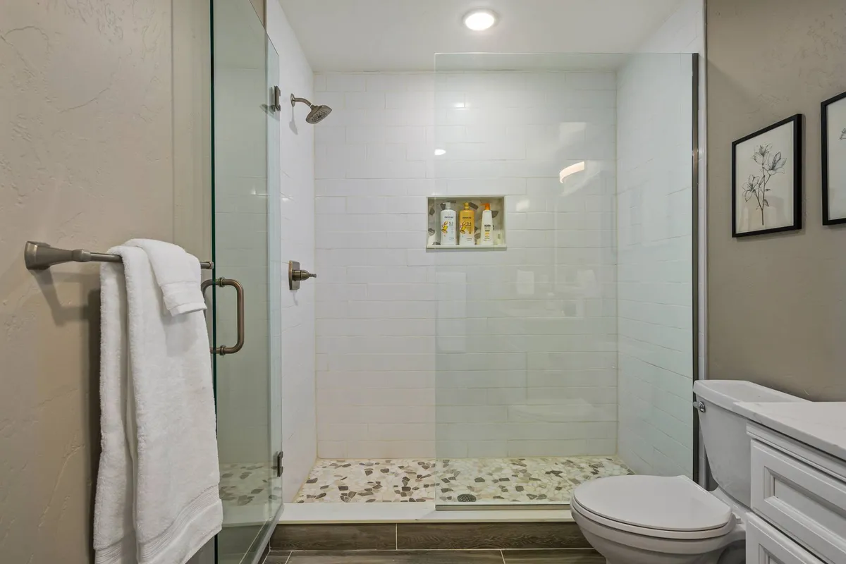 A spa-inspired bathroom featuring a glass shower enclosure with a pebble stone floor, shelf with toiletries, and framed botanical prints. The space feels fresh, serene, and thoughtfully designed for relaxation.
