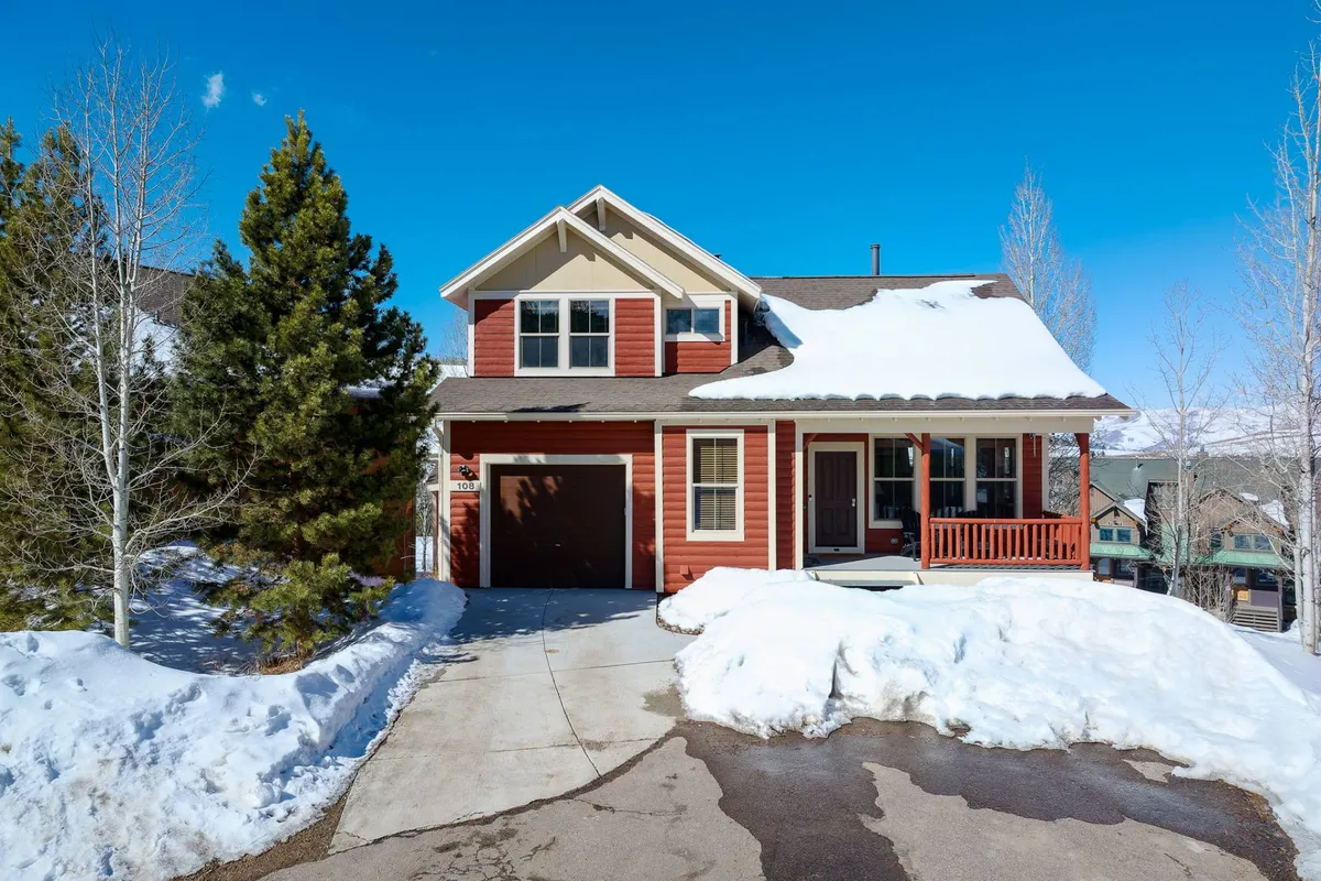 This image showcases the inviting red exterior of Timber Court surrounded by a snow-covered driveway and green trees. The porch features rocking chairs, suggesting a welcoming space for gathering and relaxing after outdoor adventures.